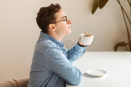 Photo for Relaxed caucasian young man student with curly hair and glasses enjoying a moment of reflection with a hot cup of coffee in a serene cafe environment. Spare time, rest, relax - Royalty Free Image