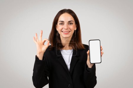 Foto de Alegre mujer de negocios caucásica milenaria mostrando una pantalla de teléfono inteligente en blanco y haciendo una señal de aprobación, lo que indica la aprobación o una revisión positiva de una aplicación móvil o servicio - Imagen libre de derechos