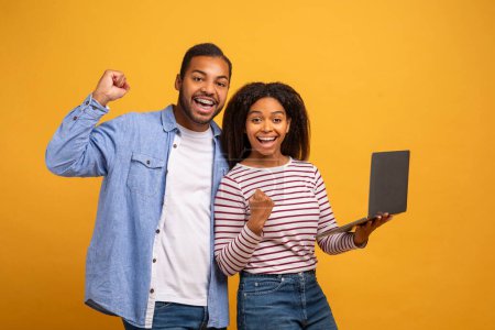 Téléchargez les photos : Gagner en ligne. Joyeux couple afro-américain avec ordinateur portable célébrant le succès, jeune homme et femme noirs heureux regardant l'écran d'ordinateur et secouant les poings, debout sur fond jaune, Copier l'espace - en image libre de droit
