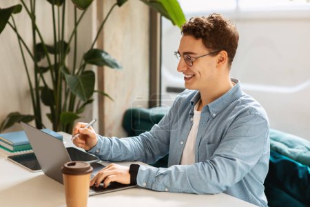 Photo for Content caucasian young man student with curly hair, wearing glasses, happily working on his tablet with a stylus and a coffee cup nearby in a plant-filled cozy workspace - Royalty Free Image