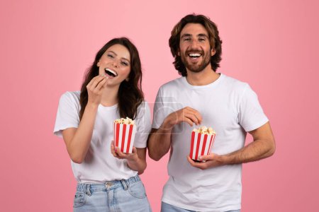 Foto de Alegre alegre europeo mujer y hombre en blanco camisetas riendo y comiendo palomitas de maíz de rojo y blanco rayas cajas, disfrutando de un divertido momento juntos en un rosa fondo, estudio - Imagen libre de derechos
