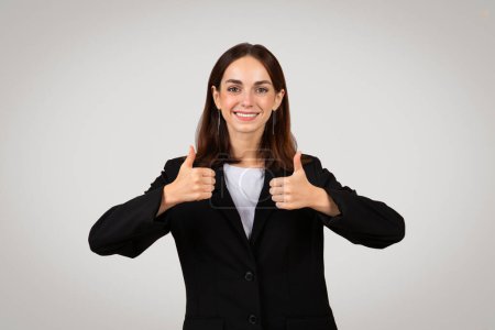 Foto de Sonriente joven mujer de negocios caucásica en un traje negro dando pulgares hacia arriba con ambas manos, expresando entusiasmo y aprobación con un comportamiento amistoso, aislado sobre fondo gris, estudio - Imagen libre de derechos