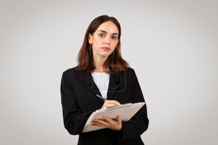 Photo for Serious millennial caucasian businesswoman in a formal black suit writing notes on a clipboard, displaying focus and professionalism in a corporate environment, isolated on gray background, studio - Royalty Free Image