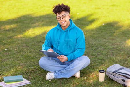 Photo for Smiling Brazilian student guy browsing internet on digital tablet outside, learning at city park on green lawn, studying with workbooks and backpack, looks at camera with smile - Royalty Free Image