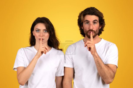Téléchargez les photos : Une jeune femme européenne sérieuse et un homme aux cheveux foncés et aux t-shirts blancs geste de silence, en plaçant leurs doigts sur leurs lèvres, sur un fond jaune vif, studio - en image libre de droit