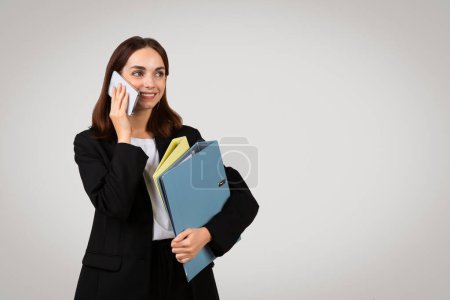 Foto de Comprometida y optimista millennial mujer de negocios caucásica conversando en un teléfono inteligente mientras sostiene carpetas coloridas, mostrando multitarea y comunicación efectiva en un entorno profesional - Imagen libre de derechos