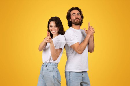 Selbstbewusstes kaukasisches junges Paar in weißen T-Shirts, Rücken an Rücken stehend, Daumen hoch mit verspielter Haltung auf sonnigem gelbem Hintergrund, Studio. Spiel, Spaß in der Freizeit