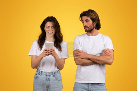 Téléchargez les photos : Femme caucasienne millénaire souriante joyeusement absorbée dans son smartphone avec un homme curieux à côté d'elle, bras croisés, tous deux portant des t-shirts blancs décontractés sur fond jaune, studio - en image libre de droit