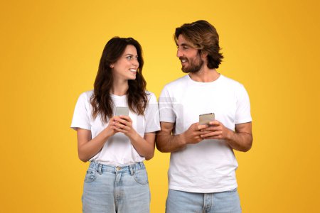 Photo for Happy millennial caucasian couple engaging with each other, holding smartphones and smiling, woman looking at man with interest, both in white shirts on a yellow background, studio - Royalty Free Image