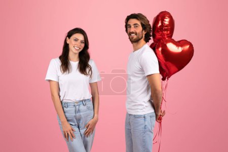 Photo for Smiling european woman and smiling man in white t-shirts, with the man holding a bunch of red heart-shaped balloons, celebrating a romantic occasion on a pink background, studio - Royalty Free Image