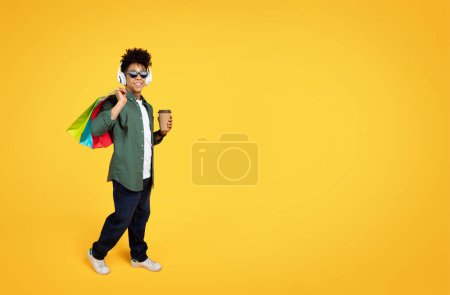 Foto de Cool joven afroamericano elegante con gafas de sol y auriculares inalámbricos escuchando música mientras va de compras, llevando coloridas bolsas de papel y café para llevar, fondo amarillo, espacio para copiar - Imagen libre de derechos