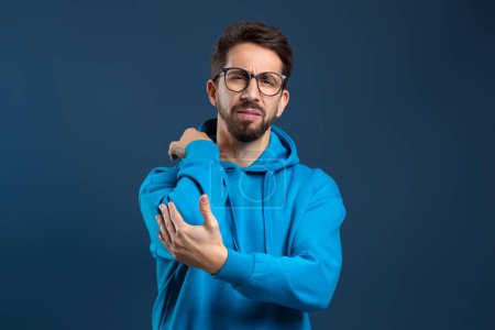 Young upset man clutching his elbow in pain or discomfort, millennial guy feeling unwell, indicating an injury or trauma, injured male standing against dark blue studio background, copy space