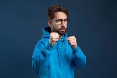 Determined man wearing eyeglasses and blue hoodie raising his fists in defensive pose, confident serious male looking ready for challenge with focused look, standing against dark studio background