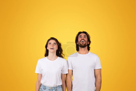 Téléchargez les photos : Excité choqué heureux millénium caucasien homme et femme en chemises blanches levant les yeux dans la crainte, leurs expressions pleines de joie et d'émerveillement sur un fond jaune uni, studio - en image libre de droit