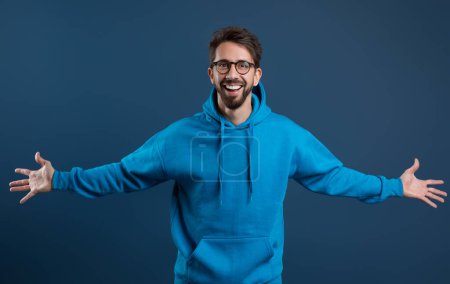 Photo for Cheerful Young Guy In Blue Hoodie Spreading Arms And Smiling To Camera, Handsome Happy Man Standing Against Dark Studio Background, Joyful Millennial Male Expressing Positivity, Free Space - Royalty Free Image