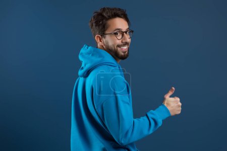 Photo for Happy Young Man Turning At Camera And Showing Thumb Up, Cheerful Millennial Guy In Eyeglasses Gesturing Sign Of Approval, Recommending Something While Standing Against Blue Studio Background - Royalty Free Image