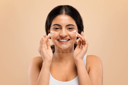 Foto de Rutina de belleza. Mujer india joven feliz aplicando crema facial blanca en sus mejillas con expresión alegre, alegre hermosa hembra haciendo cuidado diario de la piel, de pie contra fondo beige suave - Imagen libre de derechos