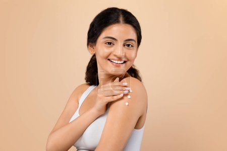 Foto de Cuidado diario de la piel. Mujer india joven con una sonrisa agradable aplicando suavemente una pizca de crema en su hombro, atractiva dama oriental ilustrando la rutina de belleza, de pie contra el fondo beige del estudio - Imagen libre de derechos