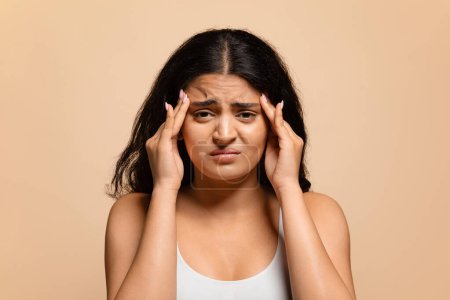 Headache Concept. Stressed indian woman in white top touching her temples, upset eastern female looking at camera with pained expression on her face, having migraine, standing on beige background