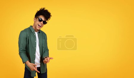Foto de Elegante chico afroamericano milenario con el pelo rizado usando ropa casual y gafas de sol haciendo gestos en el fondo del estudio amarillo, apuntando a la cámara y sonriendo, panorama con espacio para copiar - Imagen libre de derechos