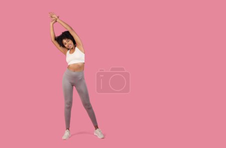Joyful young African American woman with curly hair stretching her arms, wearing white sports top and grey leggings on pink background, free space