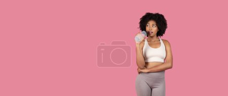 Foto de Mujer negra joven centrada en la aptitud con el pelo rizado, usando ropa deportiva, sosteniendo la botella de agua, mirando hacia otro lado en el espacio libre pensativamente sobre el fondo rosa, panorama - Imagen libre de derechos