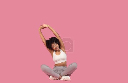 Photo for Happy African American woman in white sports top and grey leggings doing stretch with arms above head, sitting on pink background, free space - Royalty Free Image