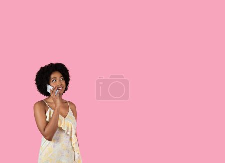Téléchargez les photos : Une jeune femme afro-américaine joyeuse en robe d'été florale parle sur un smartphone avec une expression réfléchie, regarde l'espace libre, isolée sur un fond rose, studio - en image libre de droit
