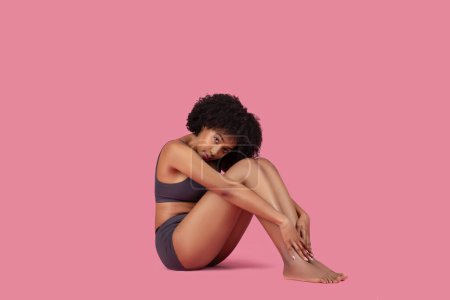 Photo for Pensive young African American woman in sportswear or underwear sitting and touching her foot on pink background, looking at camera, free space - Royalty Free Image