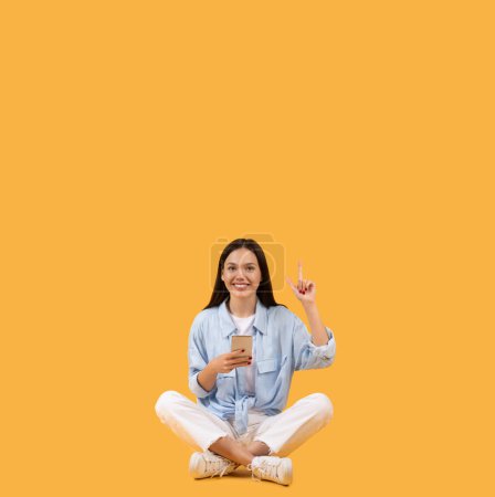 Photo for Cheerful young woman sits cross-legged on yellow background, holding smartphone and pointing upwards at free space with look of inspiration and joy - Royalty Free Image