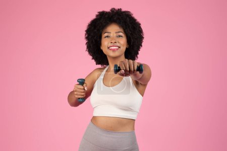 Téléchargez les photos : Radieuse jeune femme noire aux cheveux bouclés souriant tout en soulevant des poids, s'engageant dans un entraînement d'haltère, posant sur fond de studio rose - en image libre de droit