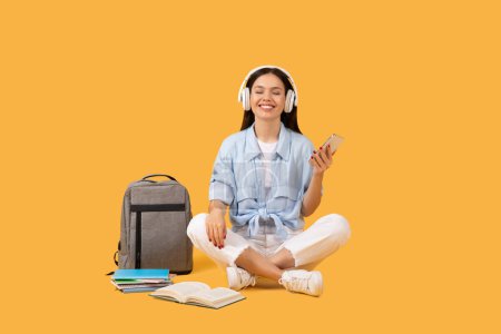 Photo for Content lady student takes study break, listening to music with headphones and holding smartphone, with books and backpack suggesting learning environment - Royalty Free Image