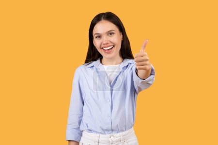Téléchargez les photos : Jeune femme souriante en chemise bleue décontractée donne pouces vers le haut signe, exprimant la positivité et l'approbation, debout sur fond jaune vibrant - en image libre de droit