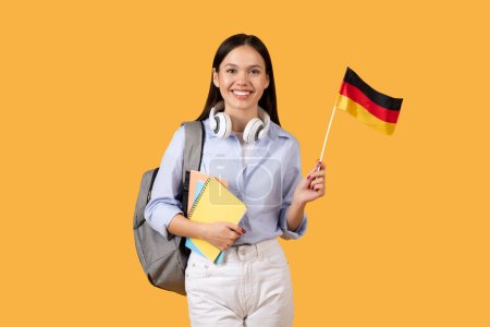 Fröhliche Studentin mit Kopfhörern um den Hals hält deutsche Flagge und bunte Notizbücher in der Hand, genießt das Sprachenlernen, posiert vor leuchtend gelbem Hintergrund