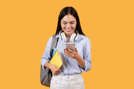 Téléchargez les photos : Étudiante joyeuse avec sac à dos et cahiers à la main, sourit alors qu'elle écrit sur son téléphone portable, incarnant un mélange d'étude et de technologie sur fond jaune vif - en image libre de droit