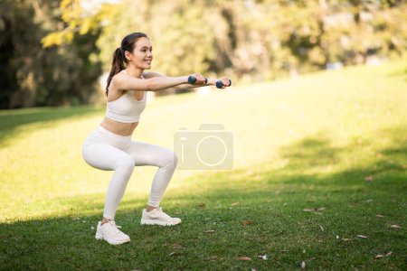 Téléchargez les photos : Jeune femme caucasienne athlétique millénaire en vêtements de sport blancs faisant squats avec haltères dans un parc vert ensoleillé, favorisant la forme physique et le mode de vie actif, à l'extérieur, pleine longueur - en image libre de droit