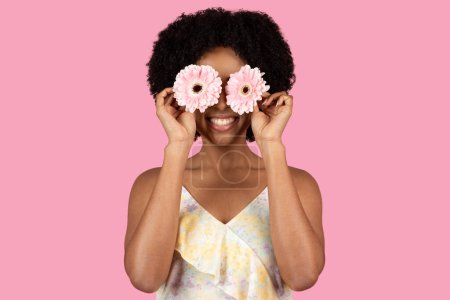 Téléchargez les photos : Délicieuse heureuse femme afro-américaine millénaire drôle utilisant deux marguerites de gerbera rose comme lunettes ludiques, avec un sourire rayonnant, sur un fond rose minimaliste, studio - en image libre de droit