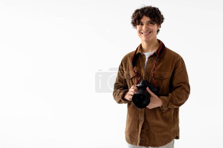 Foto de Alegre joven fotógrafo masculino con el pelo rizado, con chaqueta de pana, sostiene la cámara réflex digital profesional, listo para la sesión de fotos, fondo blanco con espacio libre - Imagen libre de derechos