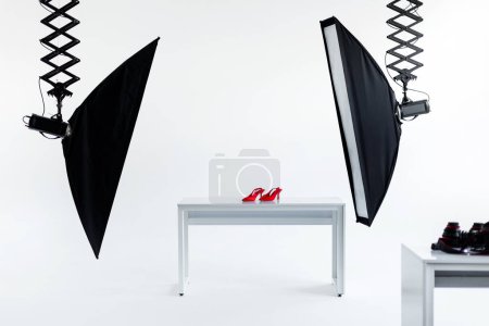 Photo for Modern photography studio setup with professional lighting equipment poised to capture vibrant red high-heeled shoes on clean white table - Royalty Free Image
