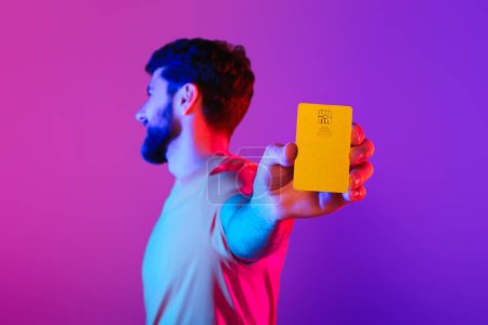 Photo for Digital Shopping. Man holding and showing credit card in purple neon studio, advertising online commerce, showcasing limitless possibilities and ease of financial transactions. Shallow depth - Royalty Free Image
