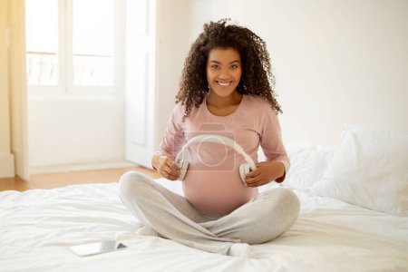 Foto de Mujer embarazada negra feliz sosteniendo auriculares cerca del vientre, tocando la melodía del bebé en el útero, madre embarazada afroamericana escuchando música clásica durante el embarazo, sentada en la cama en casa - Imagen libre de derechos