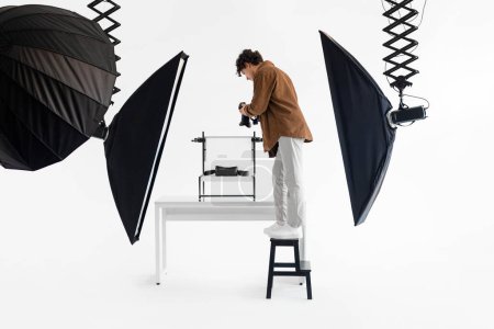 Fotógrafo masculino enfocado se para en el taburete, ajustando meticulosamente su cámara para capturar la toma de estudio perfecta, rodeado de equipo de iluminación profesional