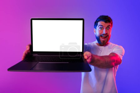 Photo for Emotional Young European guy showcasing blank laptop screen, advertising websites and online business concepts, standing holding computer on purple blue studio backdrop. Freelance opportunities - Royalty Free Image