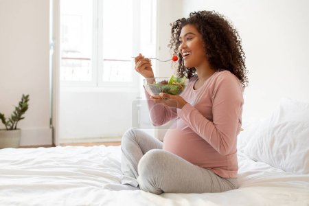 Foto de Hermosa mujer embarazada negra con sonrisa radiante comiendo ensalada de verduras frescas, mujer embarazada afroamericana feliz sentada en su cama en un dormitorio bien iluminado, disfrutando de una comida saludable durante el embarazo - Imagen libre de derechos