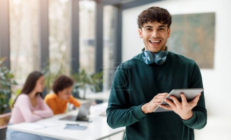 Photo for Happy young man with stylish headphones around his neck holds digital tablet, engaging with technology while his classmates work in the backdrop - Royalty Free Image