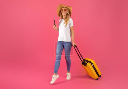Téléchargez les photos : Vibes de vacances. Femme voyageuse blonde debout avec des bagages et des billets, la promotion de la joie de voyager, posant sur fond de studio rose, la publicité des visites saisonnières et l'assurance voyage - en image libre de droit