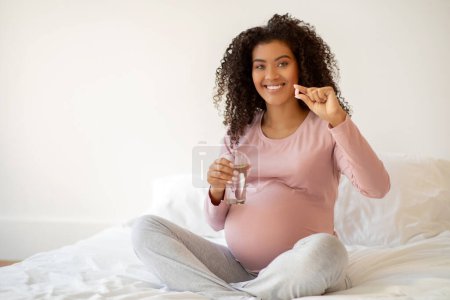 Foto de Mujer embarazada negro sonriente mostrando píldora vitamínica y sosteniendo un vaso de agua, la futura madre afroamericana promueve la salud y el cuidado prenatal mientras está sentada en la cama en el acogedor dormitorio en casa - Imagen libre de derechos