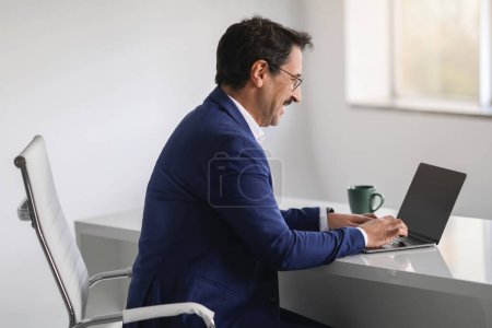 Foto de Un hombre de negocios europeo de mediana edad con un traje azul se ríe mientras escribe en una computadora portátil, posiblemente reaccionando a un momento alegre durante la comunicación remota o el trabajo. Trabajo, negocios - Imagen libre de derechos
