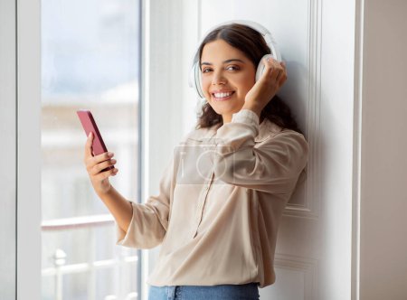 Foto de Mujer joven y feliz con auriculares inalámbricos disfrutando de escuchar música en el teléfono inteligente, sonriendo hermosa mujer de pie junto a la ventana en la habitación soleada, disfrutando de relajarse en casa, espacio libre - Imagen libre de derechos