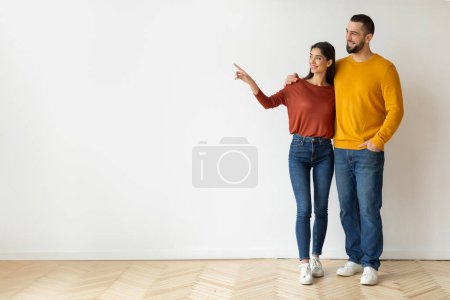 Photo for Look There. Happy caucasian couple pointing aside at copy space while standing against white wall background at home, young man and woman embracing and showing free place for your design, full length - Royalty Free Image